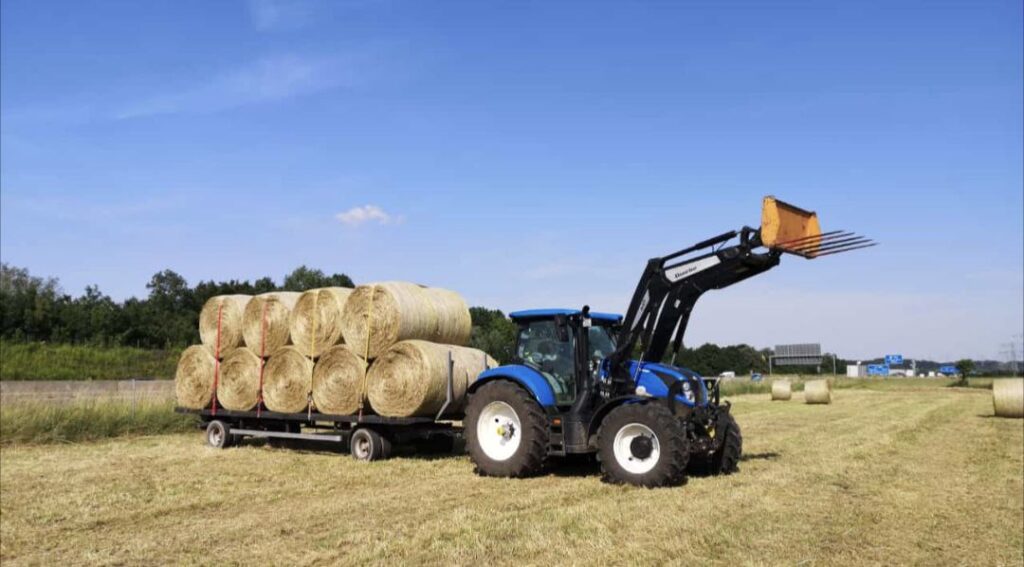 blauer Traktor mit vollbeladenem Anhänger transportiert Strohballen - nachhaltige Landwirtschaft auf dem Matzerhof in der Nähe von Augsburg in Bayern